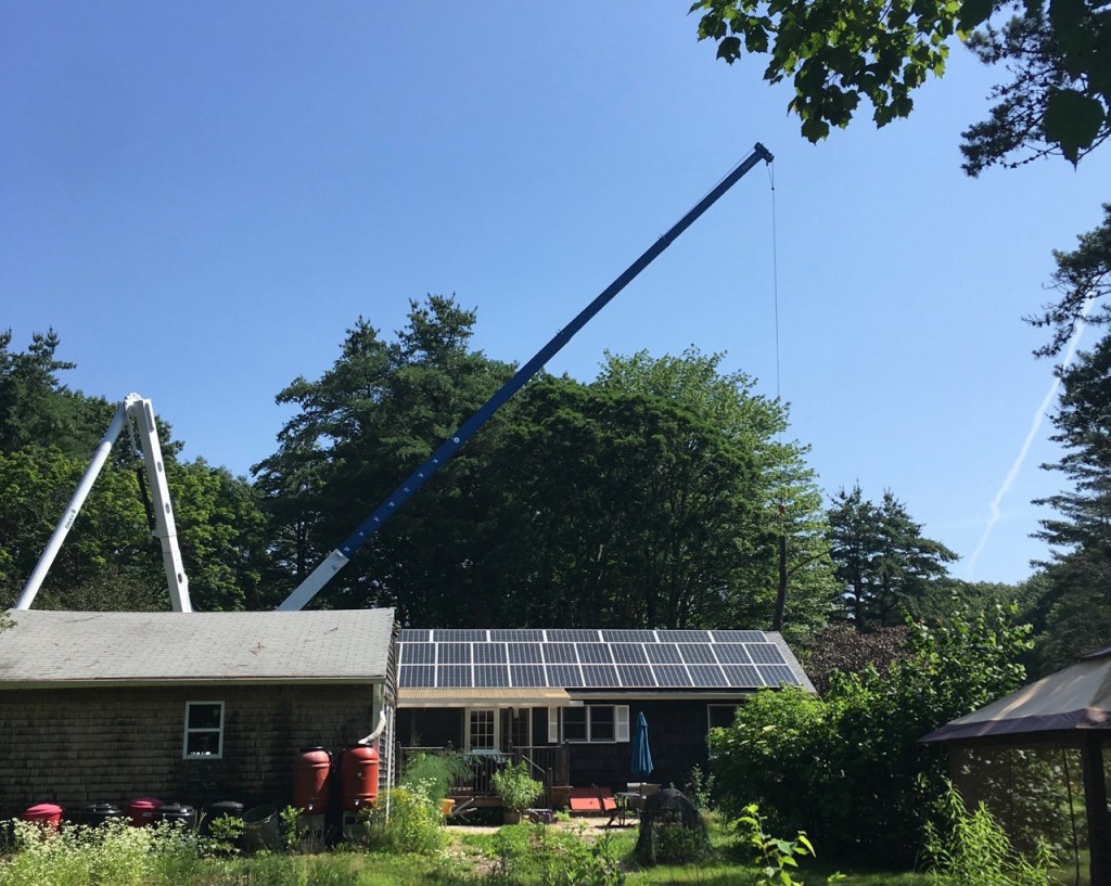 Blue sky, crane angled to the right over house with solar panels on roof, still carrying big log.