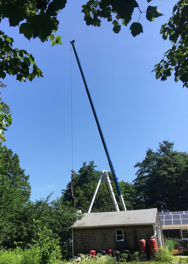 Blue sky, with tall crane and cable holding log, next to garage, trees in back.