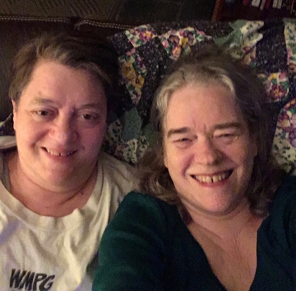 Margy & Myke selfies, two old white lesbians sitting close on a quilted background loveseat.