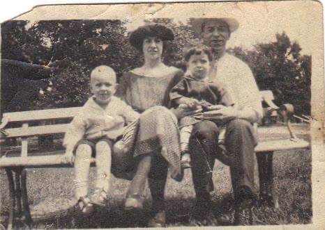 sepia toned photo of young woman and man seated on a park bench, holding two boys, one next to woman on the bench, and one on man's lap.